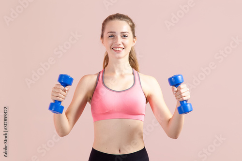 Beautiful young woman with dumbbells in her hands does fitness. Studio photoset on a pink background.