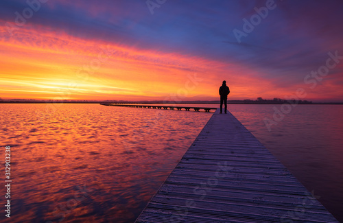 Fototapeta A man enjoying the colorful  dawn on a jetty in a lake