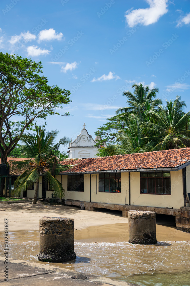 View from the pier to the residential building with a tiled roof and the shore with stone barriers. The white church in the background. Unawatuna, Sri Lanka
