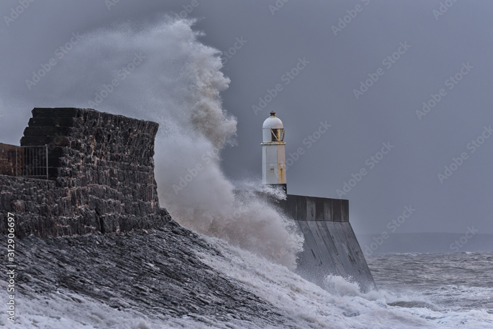 Huge waves breaking against a stone jetty, with a single lighthouse at the end, during a major winter storm.  The location is Porthcawl, on the South Wales coast.