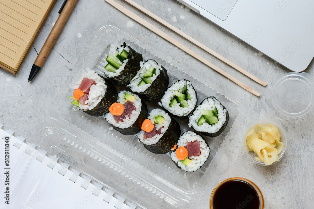 Food delivery. lunch box with sushi roll with tuna, avocado, cucumber and soy sauce on a gray office desk. takeaway lunch at the office. sushi menu delivery. Selective focus Food delivery. lunch box with sushi roll with tuna, avocado, cucumber and soy sauce on a gray office desk. takeaway lunch at the office. sushi menu delivery. Selective focus