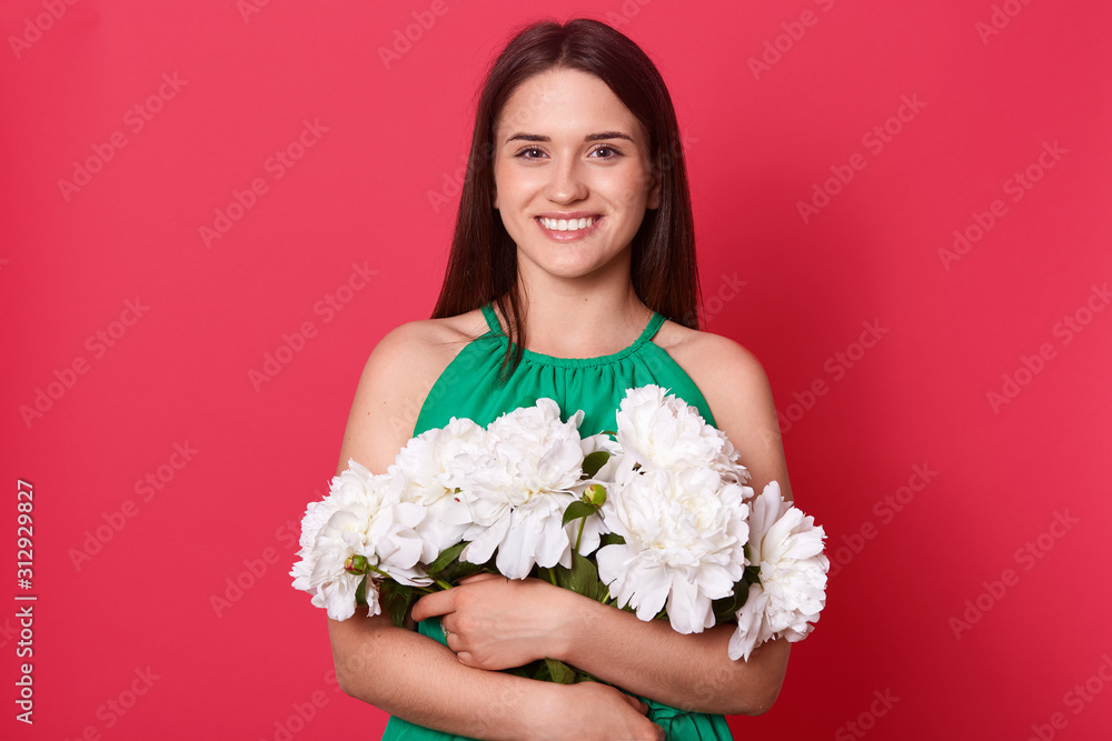Horizontal studio shot of cute cheerful positive young brunette looking directly at camera, holding bouquet of white flowers, standing isolated over red background in studio. Present concept.