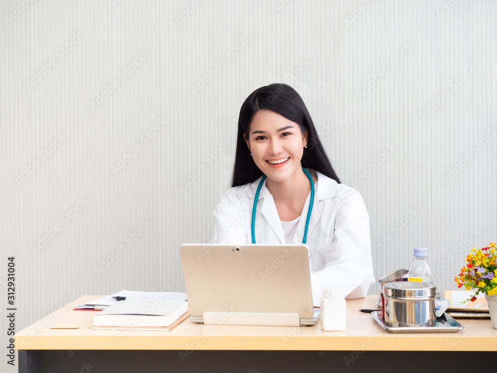 Portrait of a female doctor smiling and working with laptop on desk in the examination room. Asian beautiful doctor while working on desk in hospital or clinic. Portrait of a female doctor smiling and working with laptop on desk in the examination room. Asian beautiful doctor while working on desk in hospital or clinic.