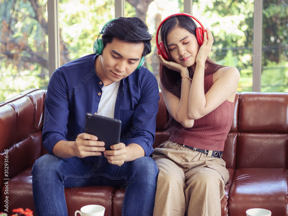 young Asian couple sitting in living room at home listen to the music from tablet , a man pick a song while woman close her eyes and listen to music happily.