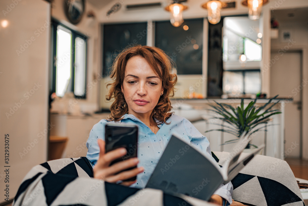 Fototapeta premium Portrait of a woman reading a text message on smart phone while reading a book at home.