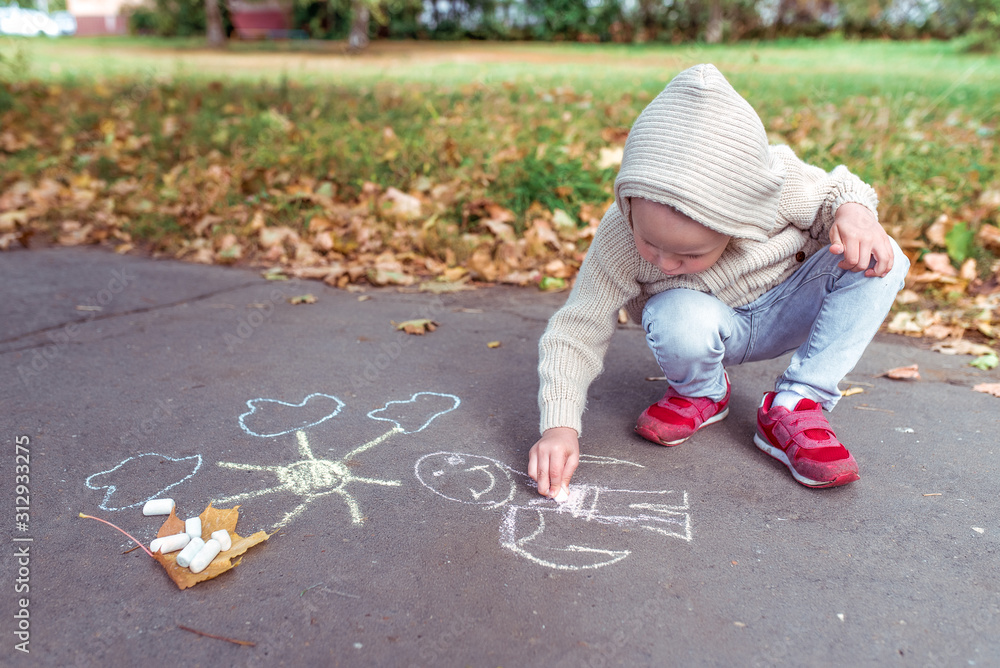 Naklejka premium Little boy 3-5 years old, draws with crayons on asphalt drawing, creative and idea art. Resting weekend, walk in autumn park. Free space for copy text. Warm casual wear, beige sweater with hood.