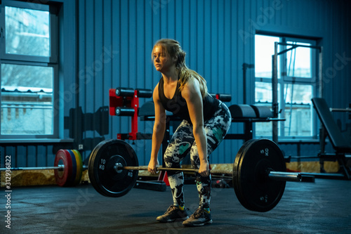 Caucasian teenage girl practicing in weightlifting in gym. Female sportive model training with barbell, looks concentrated and confident. Body building, healthy lifestyle, movement and action concept.