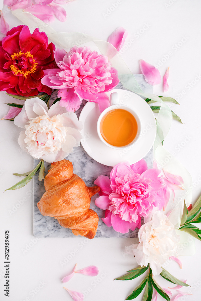 Breakfast for Valentines day with cup of tea, peony flowers and croissant on gray murble table and white background from above, flat lay, wedding, birthday, romance concept