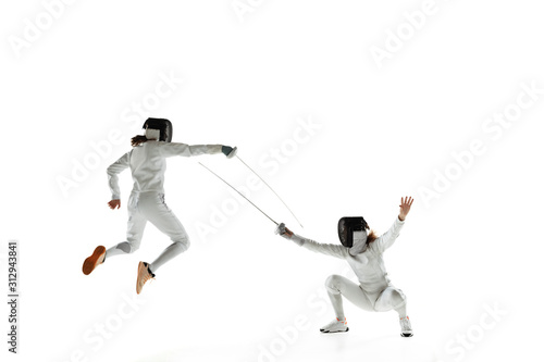 Teen girls in fencing costumes with swords in hands isolated on white studio background. Young female models practicing and training in motion, action. Copyspace. Sport, youth, healthy lifestyle.