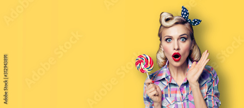 Excited surprised woman with candy lollipop. Girl pin up, gesturing. Retro fashion and vintage concept. Yellow color background. Copy space for some advertise slogan or text.