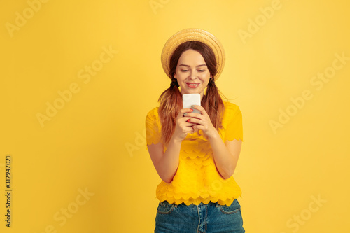 Happy using smartphone. Caucasian woman's portrait on yellow studio background. Beautiful female model in hat. Concept of human emotions, facial expression, sales, ad. Summertime, travel, resort.