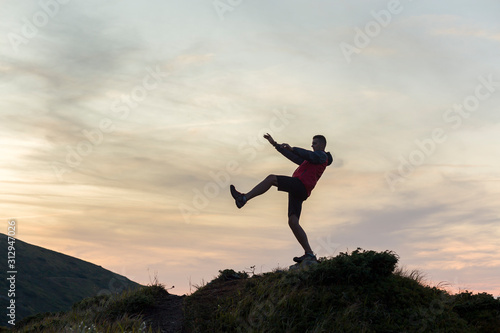 Dark silhouette of a hiker balancing on a summit stone in evening mountains.