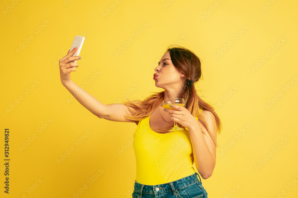 Making selfie, vlog, smiling. Caucasian woman's portrait on yellow studio background. Beautiful female model. Concept of human emotions, facial expression, sales, ad. Summertime, travel, resort. Making selfie, vlog, smiling. Caucasian woman's portrait on yellow studio background. Beautiful female model. Concept of human emotions, facial expression, sales, ad. Summertime, travel, resort.