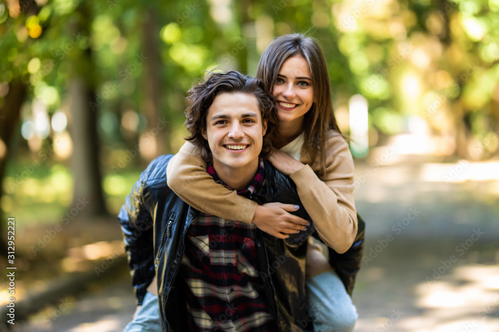 Man with stubble and headphones on neck carrying his beautiful woman in cap on back having journey trip, trendy stylish joyful couple hugging having meeting outdoors, sunny day Man with stubble and headphones on neck carrying his beautiful woman in cap on back having journey trip, trendy stylish joyful couple hugging having meeting outdoors, sunny day