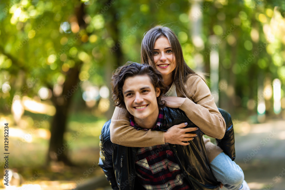 Man with stubble and headphones on neck carrying his beautiful woman in cap on back having journey trip, trendy stylish joyful couple hugging having meeting outdoors, sunny day Man with stubble and headphones on neck carrying his beautiful woman in cap on back having journey trip, trendy stylish joyful couple hugging having meeting outdoors, sunny day