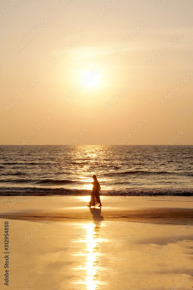 Silhouette of an unrecognizable woman in traditional Indian clothing walking on a golden sandy beach at sunset