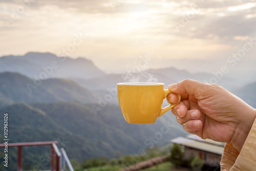 Hand holding a white cup of hot espresso coffee mugs and nature view of the mountain landscape in the morning with sunlight