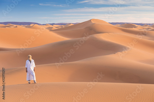 Bedouin on way through sandy desert. Beautiful sunset with big dunes on Sahara, Morocco. Silhouette nomad man. A touareg with his traditional clothes and turban. Picturesque background nature concept