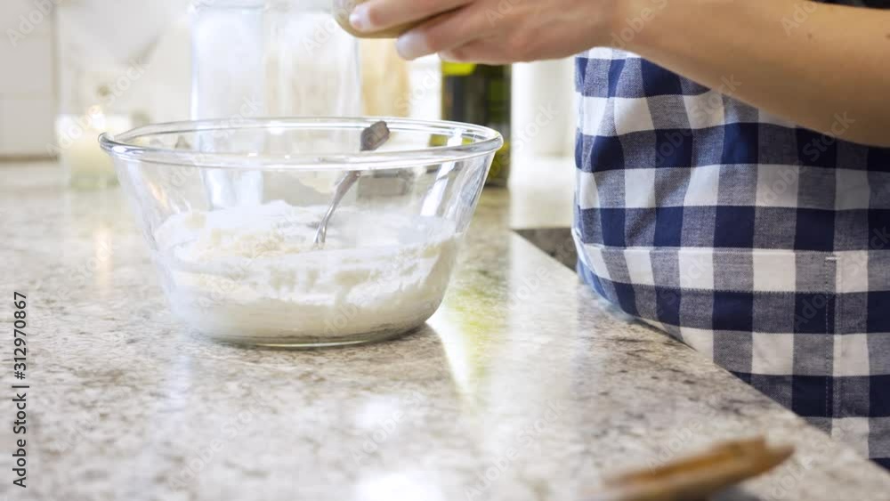 Woman preparing dough for baking bread while adding ingredients to dough and stirring in glass bowl Woman preparing dough for baking bread while adding ingredients to dough and stirring in glass bowl
