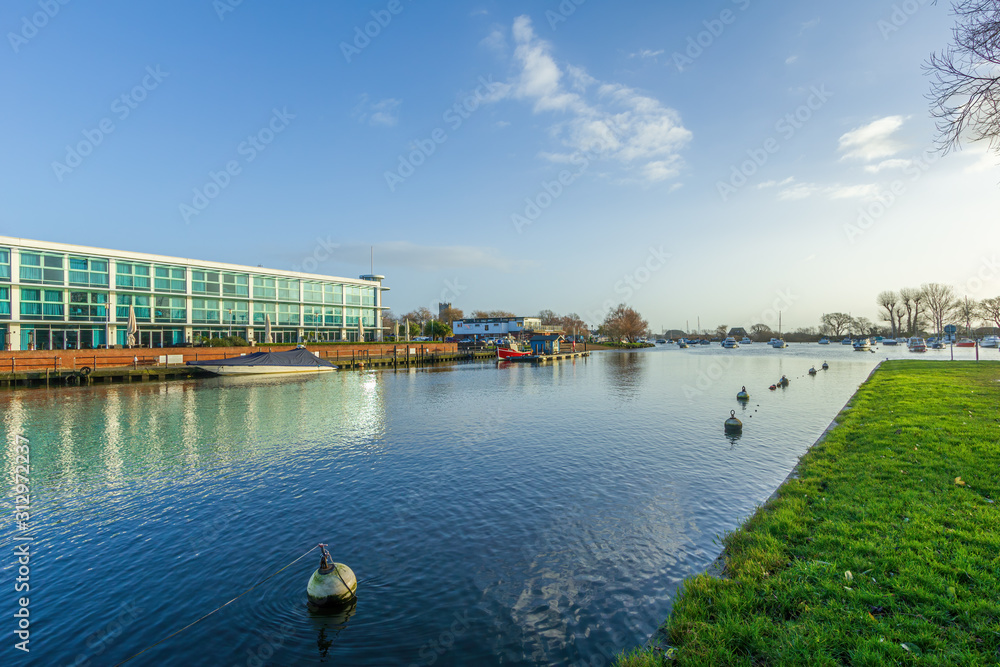 A view of a beautiful calm river with boats, grassy bank and buildings under a majestic blue sky