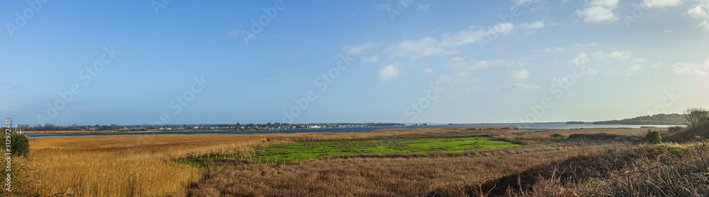 A panoramic view of the Christchurch (UK) harbor with grassy field in the foreground and houses in the background under a majestic blue sky and some white clouds