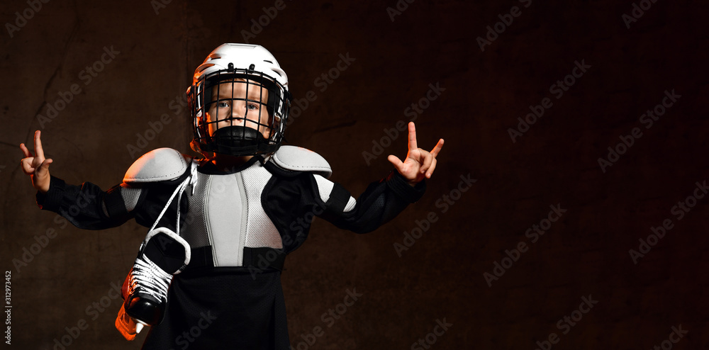 Positive boy in hockey uniform and protective helmet with hockey skates standing and showing thumb good sign