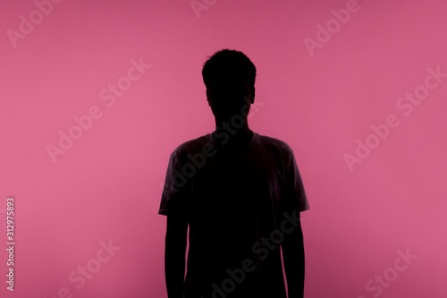 No name, anonymous person hiding face in shadow, human identity. Silhouette portrait of young man in casual T-shirt standing calm with hands down, indoor studio shot, isolated on pink background