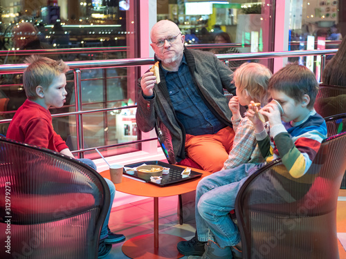 elderly pensioner in fashionable jacket with gray beard in glasses eats hamburgers at table with his grandchildren. Old hungry man bites off hamburger. Children eat and drink lemonade. Fast food cafe