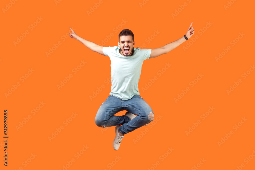 Full length of crazy overjoyed brunette man in white outfit jumping in air with raised hands, screaming loud for joy, feeling energetic and lively. indoor studio shot isolated on orange background