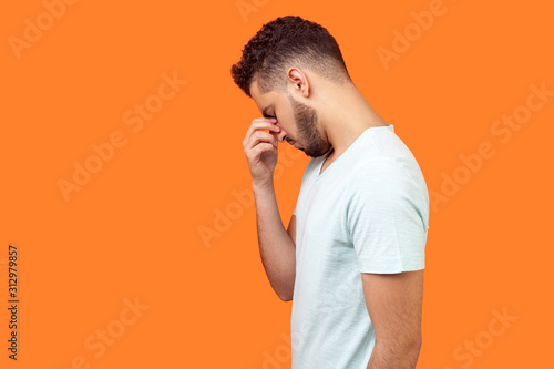 Side view of depressed brunette man with beard in white t-shirt hiding face in hand, looking desperate, is about to cry, empty copy space on left for text. studio shot isolated on orange background
