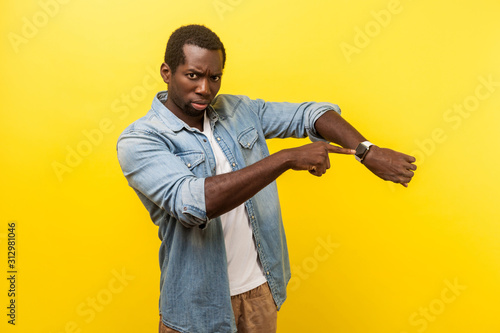Look at the time! Portrait of irritated impatient man in denim casual shirt pointing at wristwatches, nervous about being late for meeting, deadline. indoor studio shot isolated on yellow background