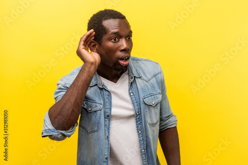 Portrait of attentive surprised man in denim casual shirt with rolled up sleeves putting hand to ear to hear better, listening carefully to rumors. indoor studio shot isolated on yellow background