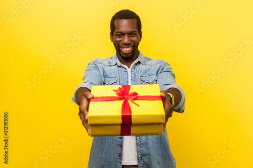 This present for you! Portrait of happy generous man in denim casual shirt giving wrapped gift box and smiling at camera, congratulating on birthday. indoor studio shot isolated on yellow background