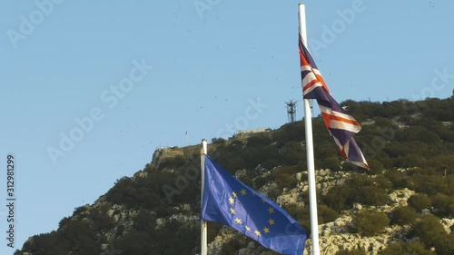 European Union flag waving along the United Kingdom's flag in the wind in Gibraltar. There are birds the background flying around the mountain.