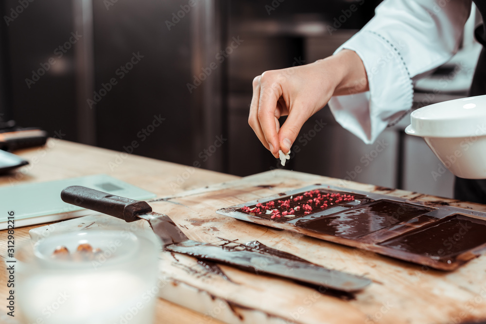 Obraz premium cropped view of chocolatier adding coconut flakes on dark chocolate bar