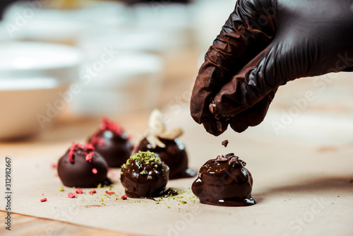 cropped view of chocolatier in black latex glove adding chocolate shavings on fresh made candies photo