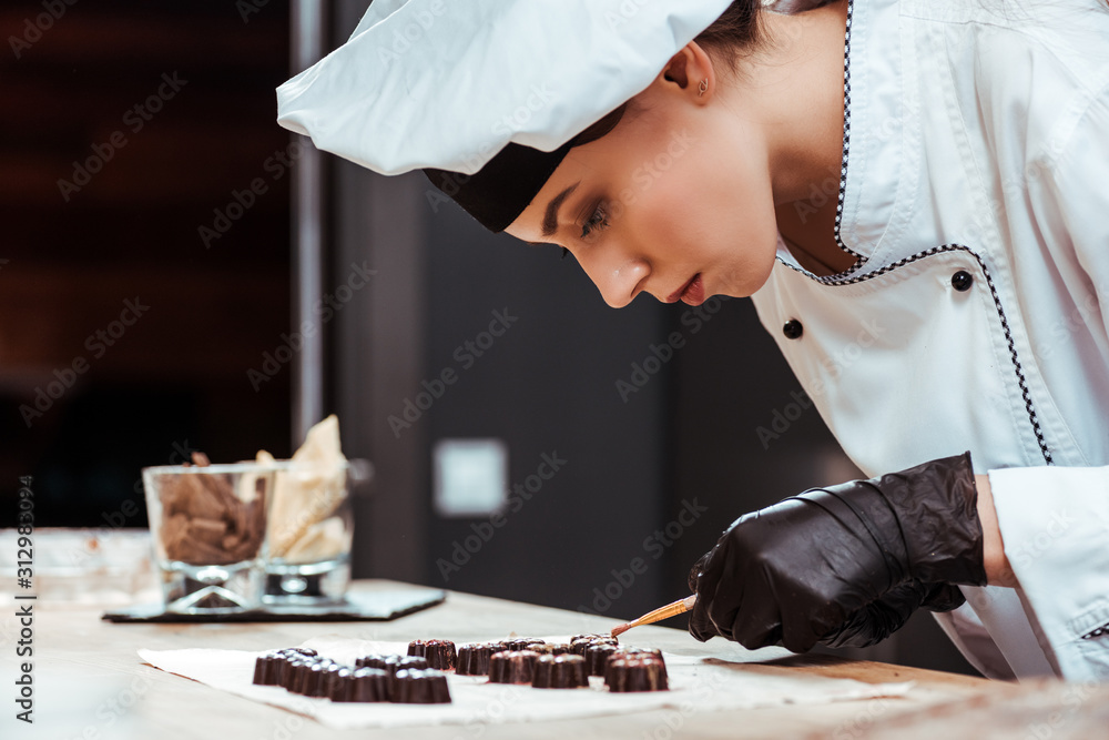 attractive chocolatier holding brush with gold powder near tasty chocolate candies on baking paper - obrazy, fototapety, plakaty 