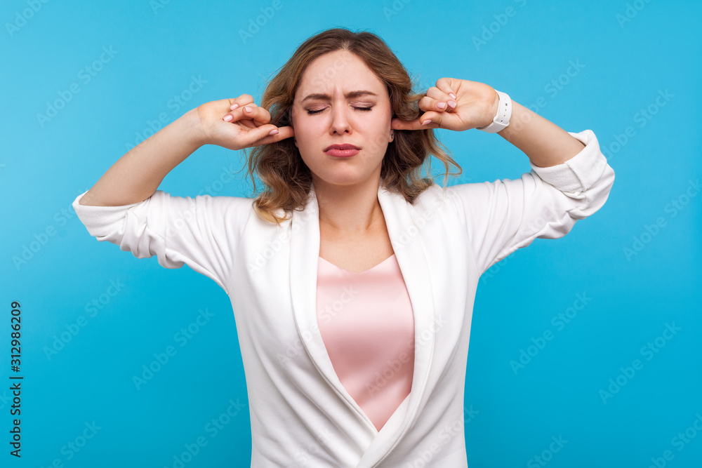 Fototapeta premium Don't want to listen! Portrait of resentful girl with wavy hair in white jacket standing covering ears and closed eyes in displeasure, frustrated about what she hear, ignoring loud noise. studio shot