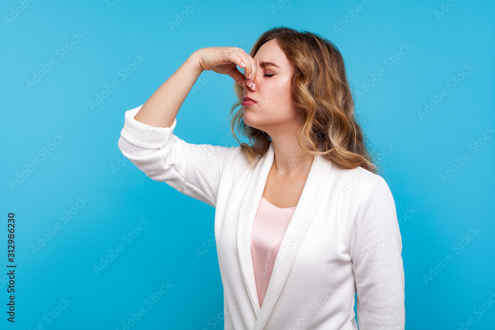 Fototapeta premium Awful smell! Portrait of woman with wavy hair in white jacket pinching her nose, expressing disgust to unpleasant odor, fart gases, her eyes closed with abhor. studio shot isolated on blue background