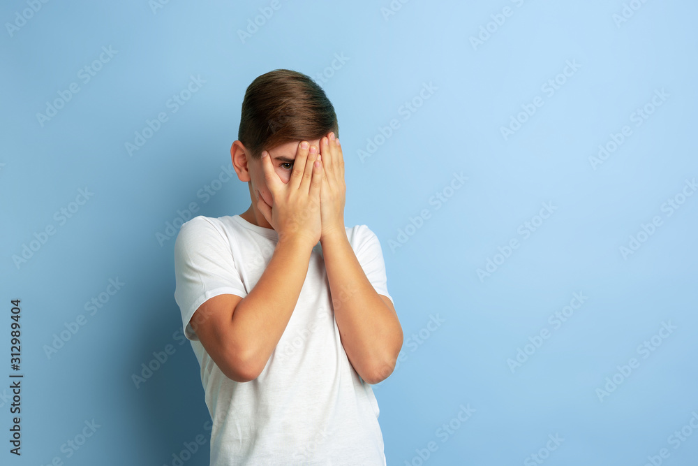 Covering face with hands. Caucasian boy portrait isolated on blue studio background. Beautiful teen male model in white shirt posing. Concept of human emotions, facial expression, sales, ad. Covering face with hands. Caucasian boy portrait isolated on blue studio background. Beautiful teen male model in white shirt posing. Concept of human emotions, facial expression, sales, ad.