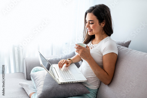 Enjoying time at home. Beautiful young smiling woman working on laptop and drinking coffee while sitting in a big comfortable sofa at home. From early morning I am checking my mailbox