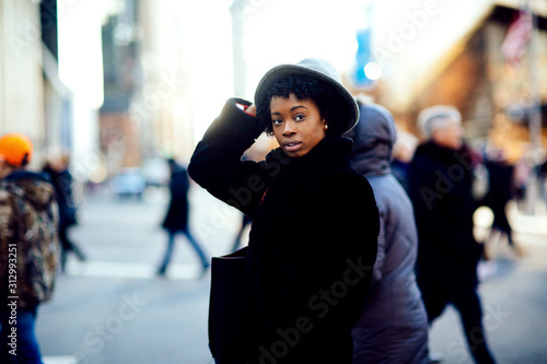 Portrait of a young woman in faux fur coat and leather bag walking on a busy big city street,holding her hat and looking at camera