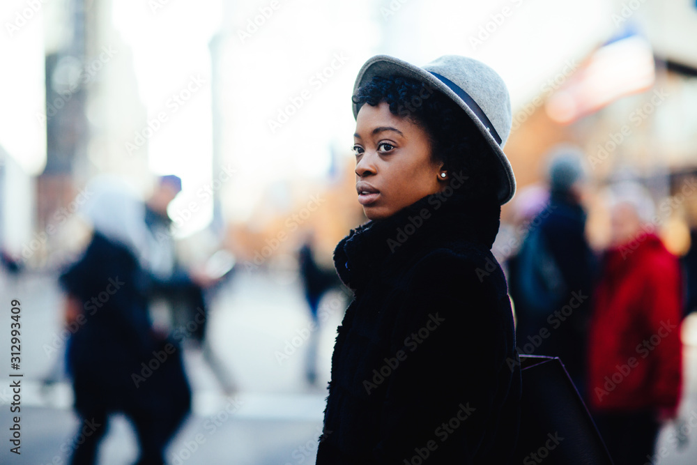 Portrait of a young woman in faux fur coat, hat and ring, walking on a busy big city street, looking up