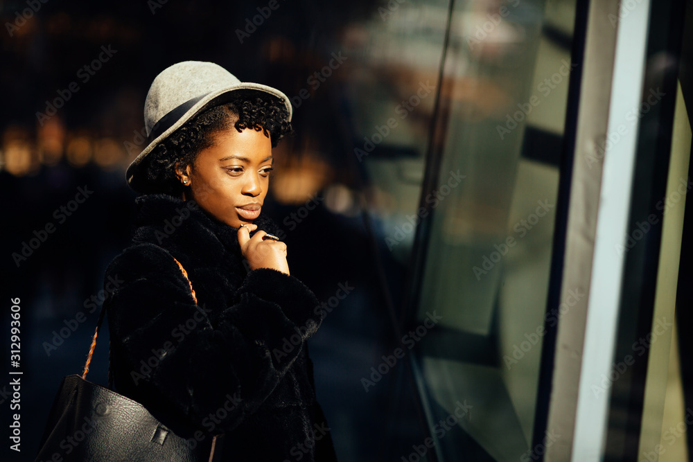 Portrait of a young woman in faux fur coat and hat standing outside in the winter sun, looking at shop window Portrait of a young woman in faux fur coat and hat standing outside in the winter sun, looking at shop window