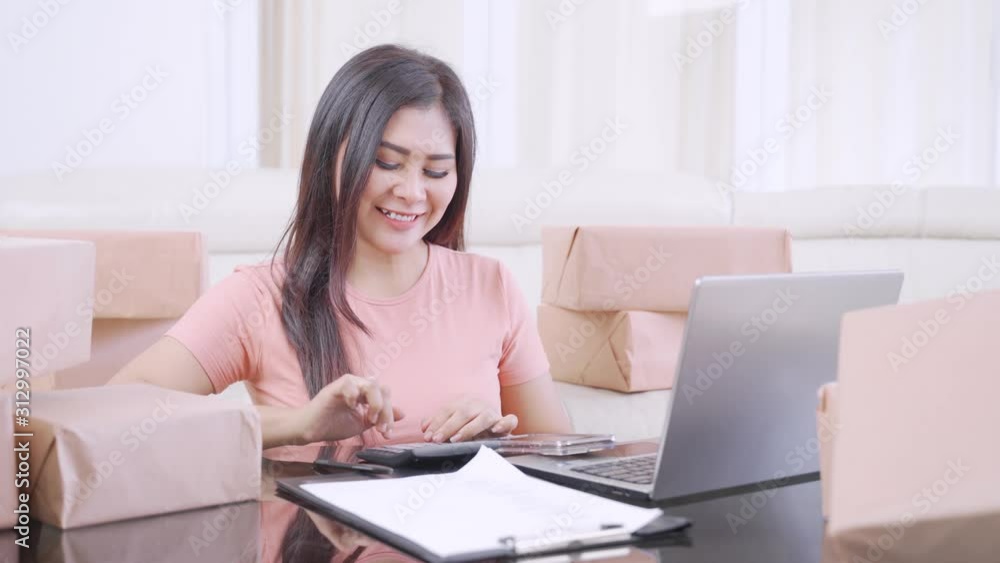Beautiful young woman working with calculator, laptop, and boxes in the living room at home. Small Medium Enterprise or Startup concept. Shot in 4k resolution Beautiful young woman working with calculator, laptop, and boxes in the living room at home. Small Medium Enterprise or Startup concept. Shot in 4k resolution