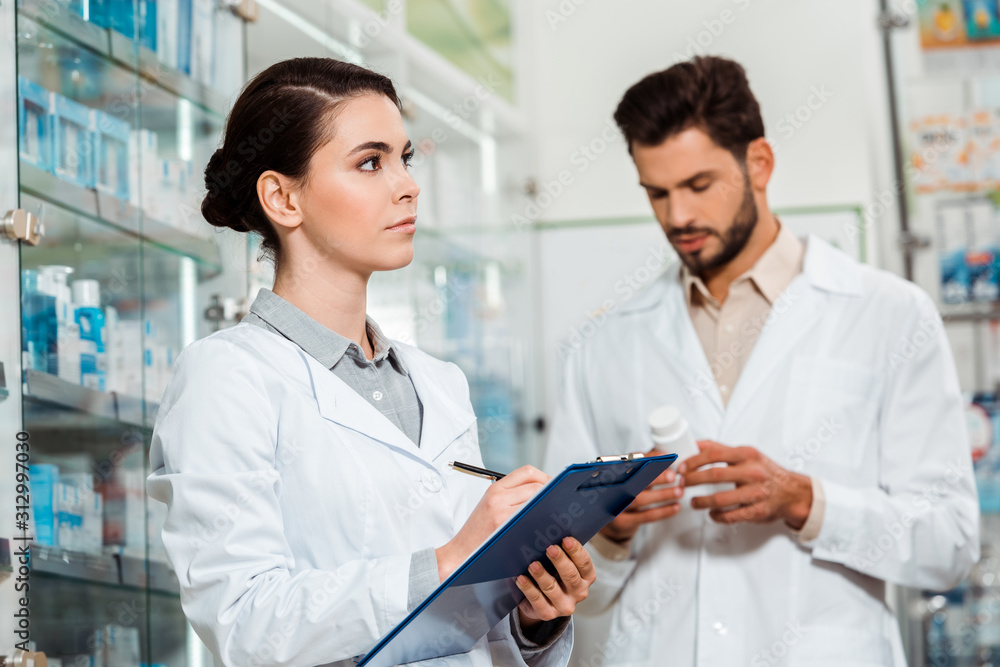 Pensive pharmacist writing on clipboard and colleague with pills on background