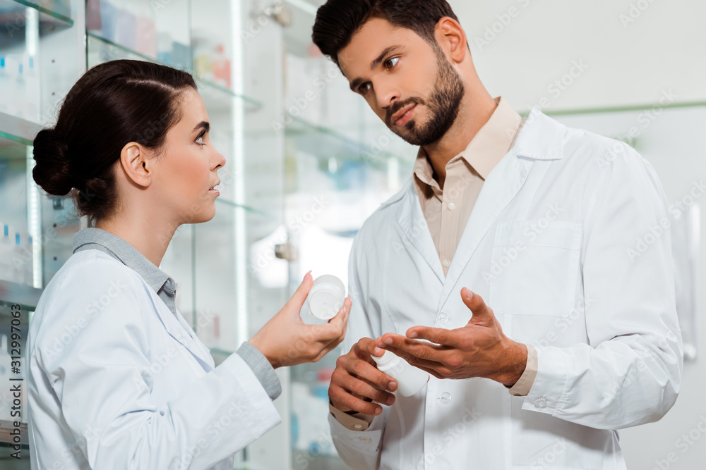Pharmacist showing to colleague jar of pills by pharmacy showcase Pharmacist showing to colleague jar of pills by pharmacy showcase