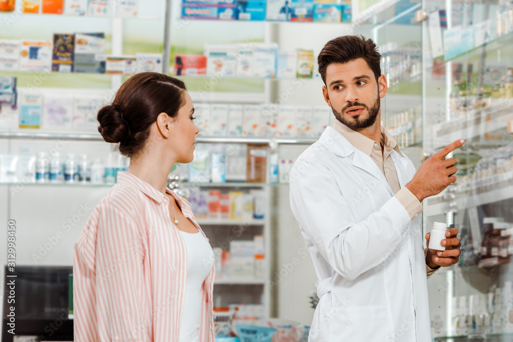 Druggist with pills pointing with finger at shelves with medicaments to customer
