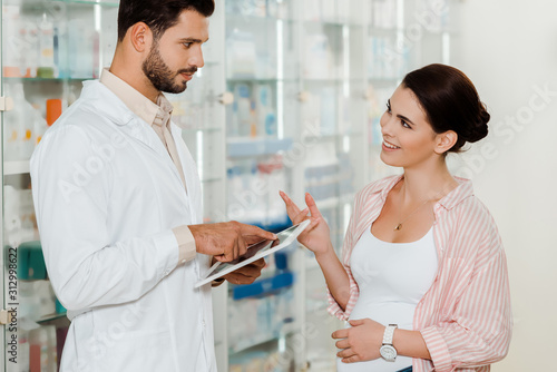 Side view of pharmacist with digital tablet looking at smiling pregnant woman in drugstore