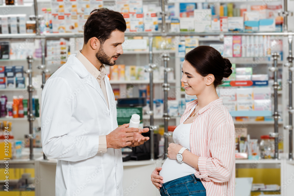 Side view of pharmacist showing jar with pills to pregnant smiling woman in apothecary Side view of pharmacist showing jar with pills to pregnant smiling woman in apothecary